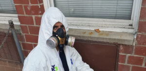 A person wearing a full white protective hazmat suit with a hood and a dual-cartridge respirator mask is looking directly at the camera. The person is situated outdoors against a red brick wall below a white window.