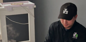 A man wearing a black shirt and a black baseball cap with a stylized white 'NYCE' logo is shown looking down, possibly working or inspecting something out of frame. There is a faint, green and black 'NYCE' logo visible on his shirt.