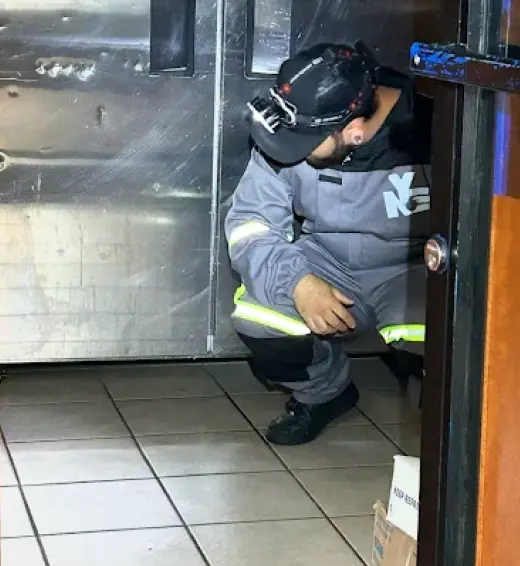 Man in grey jumpsuit with reflective stripes and a headlamp squatting near a stainless steel wall.