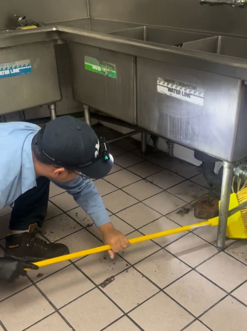 NYE technician cleaning debris from under a commercial three-compartment sink